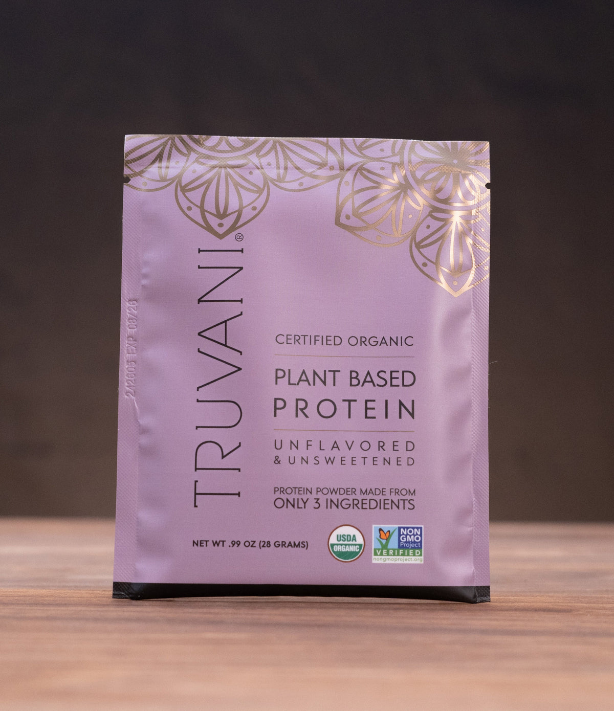 A single pouch of Truvani Certified Organic Plant Based Protein powder stands upright on a wooden surface against a dark background, with small kitchen props visible on both sides.