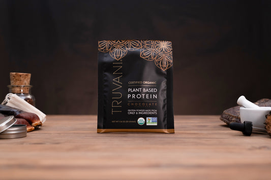 A bag of Truvani Certified Organic Plant Based Protein Chocolate flavor, standing on a wooden surface with blurred kitchen props on either side.