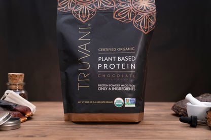 A bag of Truvani Certified Organic Plant Based Protein Powder in Chocolate flavor, displayed on a wooden surface with small kitchen props like a date palm leaf, a small bottle, and a mortar and pestle against a dark background.