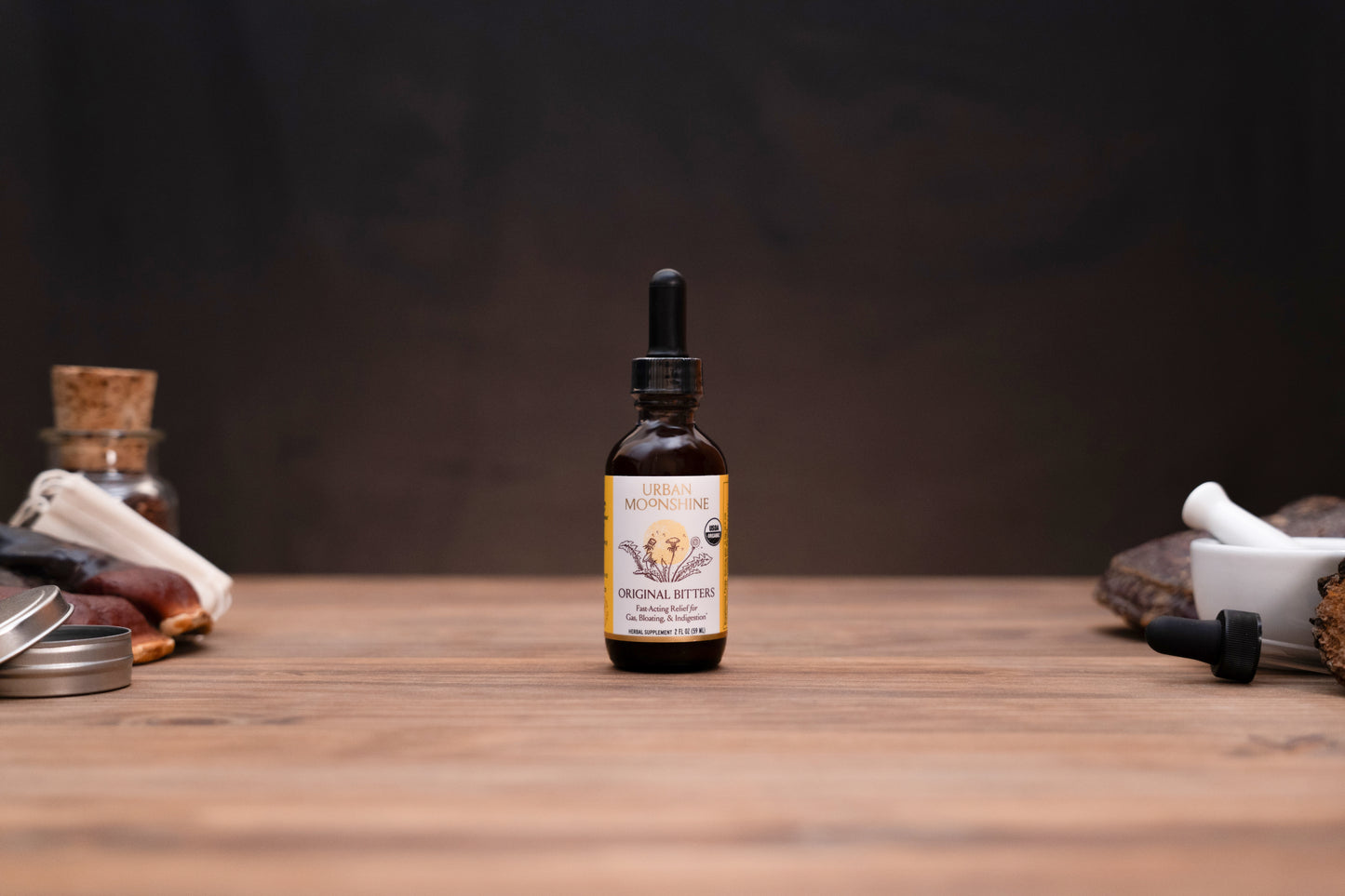 A small amber glass dropper bottle labeled Morning Glory Original Bitter stands centered on a wooden tabletop against a dark brown background, with blurred herbal props on both sides.