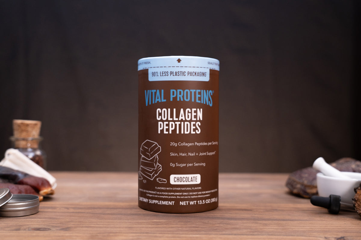A cylindrical container of Vital Proteins Collagen Peptides in Chocolate flavor, displayed on a wooden table with a dark, rustic background and small herbal props.