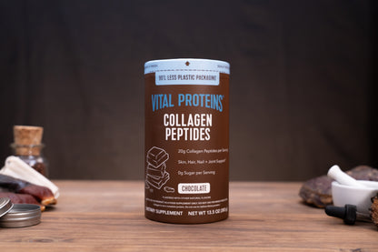 A cylindrical container of Vital Proteins Collagen Peptides in Chocolate flavor, displayed on a wooden table with a dark, rustic background and small herbal props.