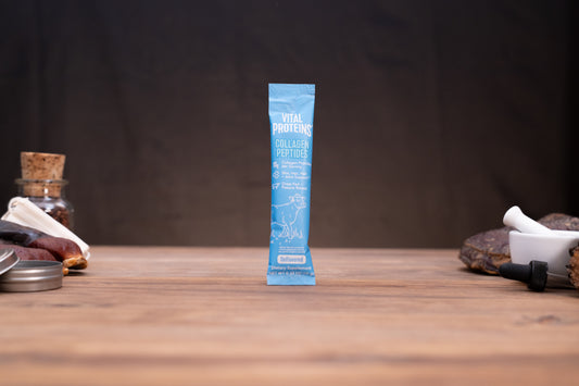 A single blue VITAL PROTEINS collagen powder stick pack stands upright on a wooden tabletop against a dark brown backdrop, with small styled props visible on both sides.