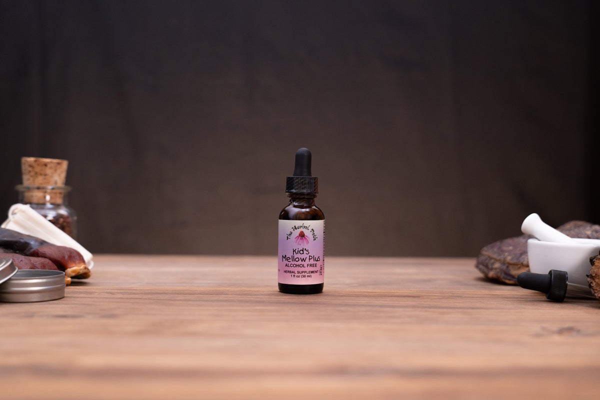 A small amber glass dropper bottle labeled 'Kiki's Medicago Hair' stands centered on a wooden tabletop, with blurred herbal props and a dark backdrop around it.