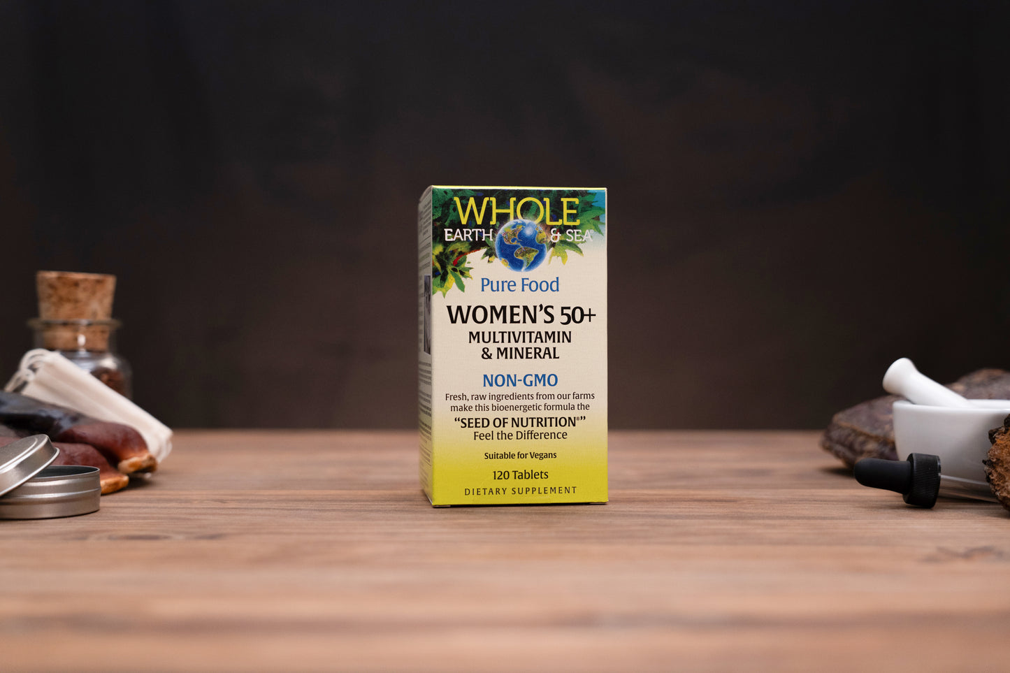 A box of Whole Earth Foods Women's 50+ Multivitamin & Mineral dietary supplement, standing on a wooden surface with rustic herbal props in the background.
