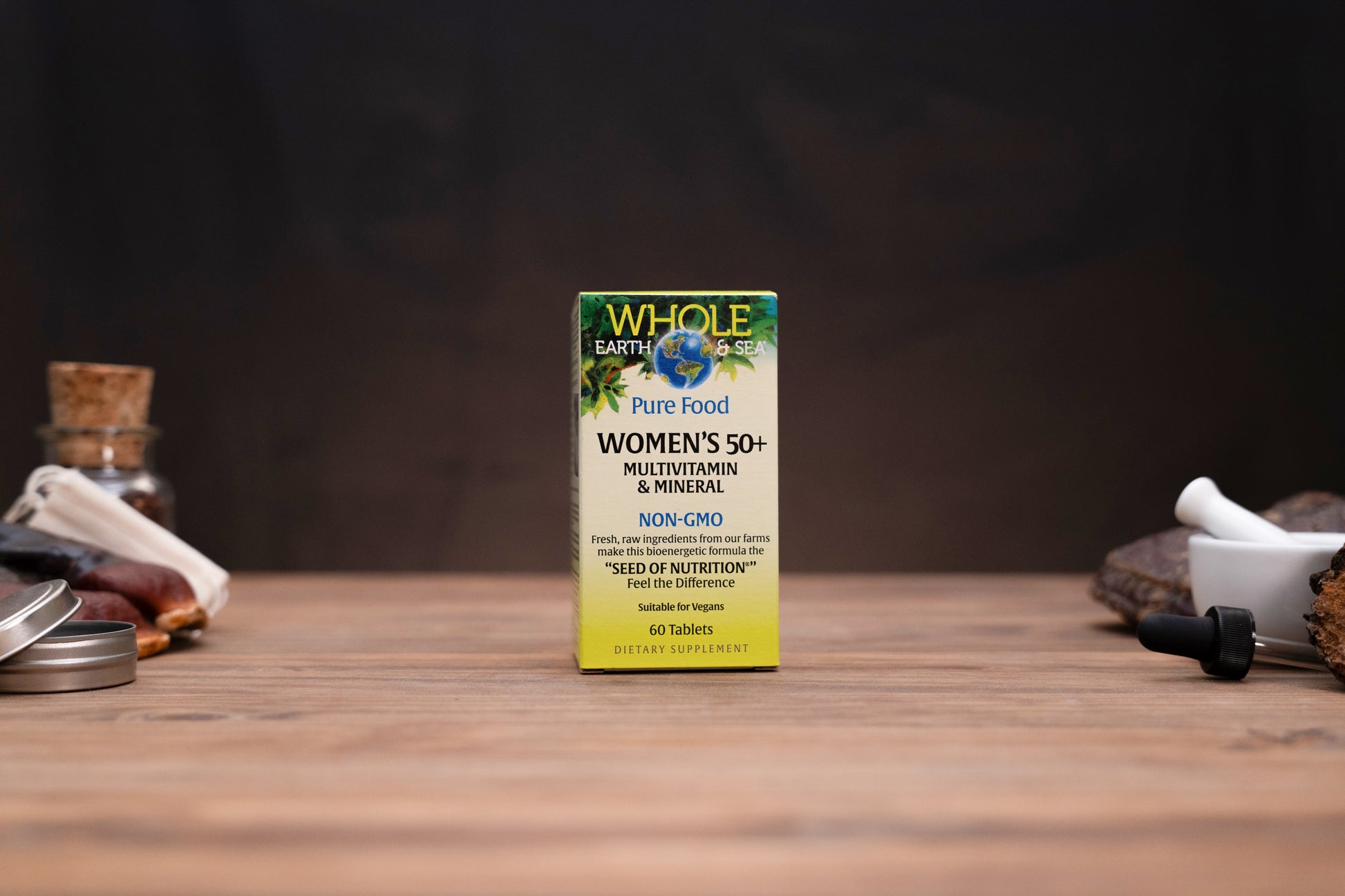 A box of Whole Earth & Me Women's 50+ Multivitamin & Mineral dietary supplement standing on a wooden tabletop, with blurred jars and a mortar-and-pestle in the background.