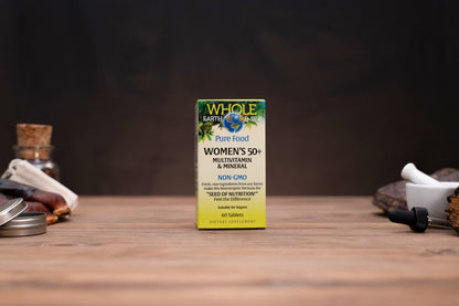 A box of Whole Earth & Me Women's 50+ Multivitamin & Mineral dietary supplement standing on a wooden tabletop, with blurred jars and a mortar-and-pestle in the background.