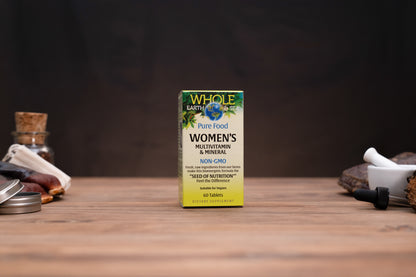 A box of Whole Earth Food Women's Multivitamin & Mineral stands on a wooden tabletop against a dark background, with small jars and a mortar-and-pestle props visible on both sides.