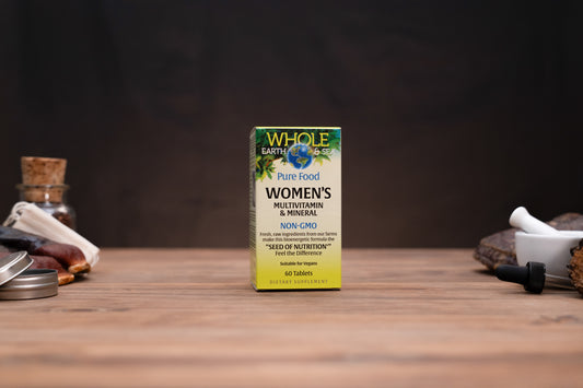 A box of Whole Earth Food Women's Multivitamin & Mineral stands on a wooden tabletop against a dark background, with small jars and a mortar-and-pestle props visible on both sides.