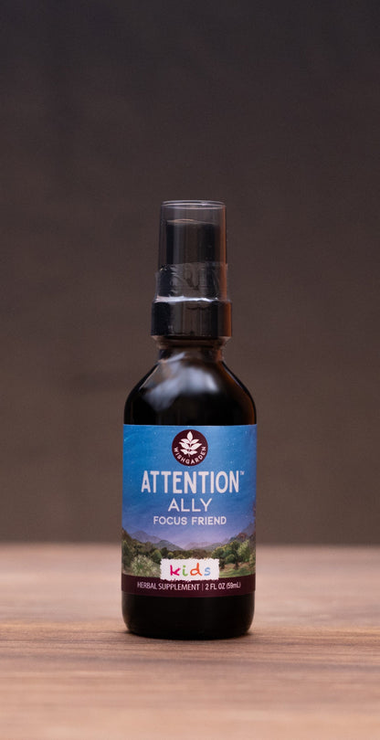 A small amber glass bottle labeled ATTENTION KILL centered on a wooden tabletop, with blurred natural props and a dark backdrop around it.