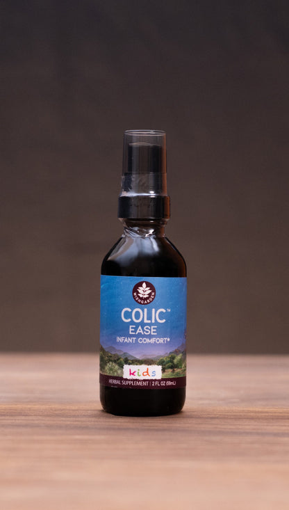 A small dark bottle labeled 'COLIC Elixir' stands centered on a wooden tabletop against a dark backdrop, with blurred herbal props on both sides.