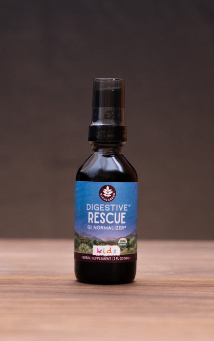A small dark bottle labeled 'COLIC Elixir' stands centered on a wooden tabletop against a dark backdrop, with blurred herbal props on both sides.