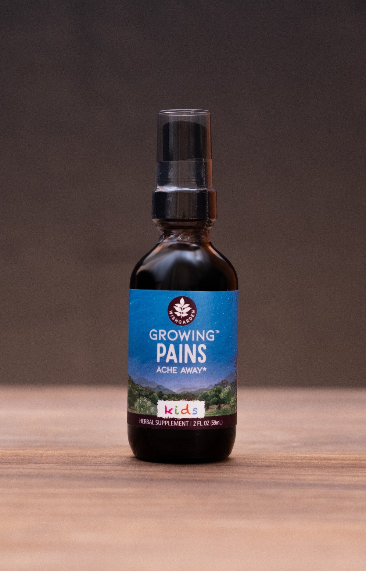 A small amber glass spray bottle with a black nozzle and a blue label reading 'GROOVY PAINS' stands centered on a wooden tabletop. The background is dark and softly blurred, with rustic props like a cork stopper, metal tin lids, and a white mortar visible at the sides.