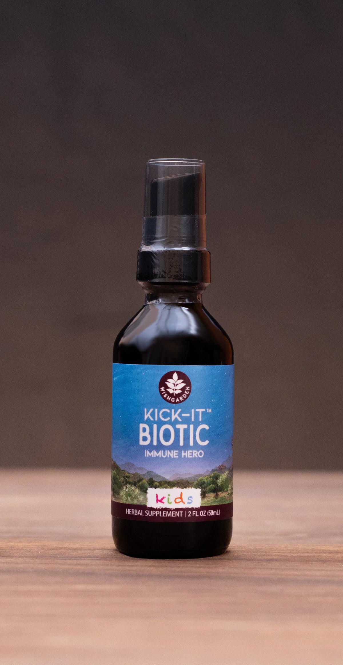 A small dark bottle labeled 'COLIC Elixir' stands centered on a wooden tabletop against a dark backdrop, with blurred herbal props on both sides.