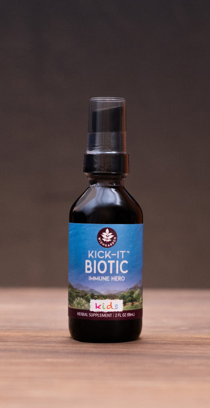 A small dark bottle labeled 'COLIC Elixir' stands centered on a wooden tabletop against a dark backdrop, with blurred herbal props on both sides.