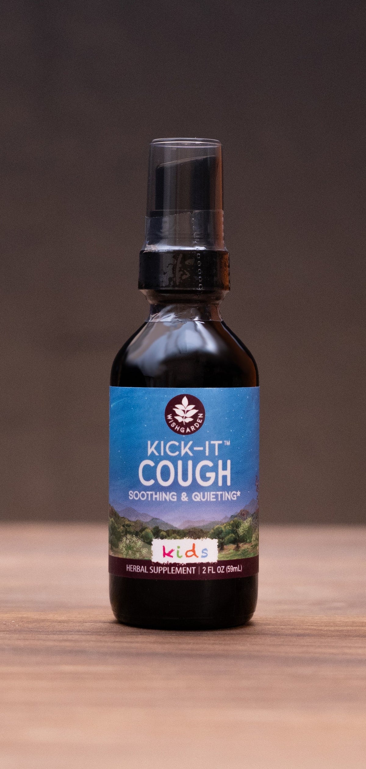 A small dark bottle labeled 'COLIC Elixir' stands centered on a wooden tabletop against a dark backdrop, with blurred herbal props on both sides.