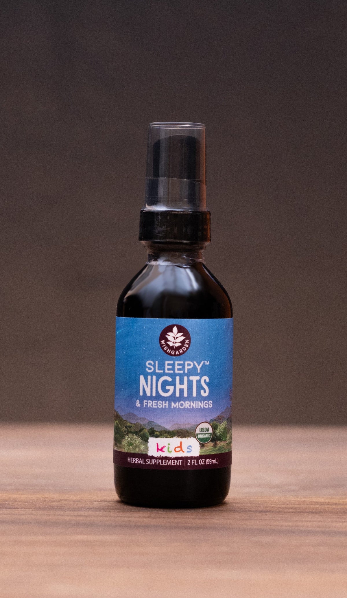 A small dark bottle labeled 'COLIC Elixir' stands centered on a wooden tabletop against a dark backdrop, with blurred herbal props on both sides.