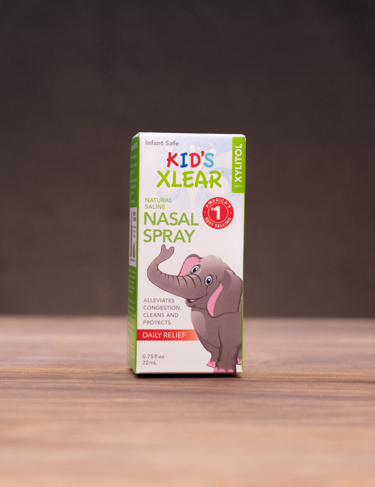 A box of Kid's Xlear 1st Nasal Spray stands centered on a wooden tabletop against a dark backdrop, with blurred props including a small bottle, stones, and a white mortar on both sides.