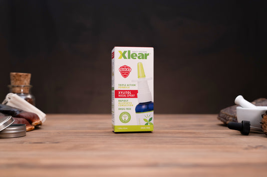 Xlear Kyltol Nasal Spray packaging box standing upright on a wooden tabletop, with blurred jars and a white dropper in the background.