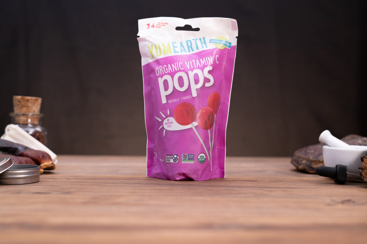 A bag of YumEarth Organic Vitamin C Pops candy stands on a wooden tabletop against a dark background, with small blurred props on both sides.
