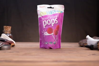 A bag of YumEarth Organic Vitamin C Pops candy stands on a wooden tabletop against a dark background, with small blurred props on both sides.