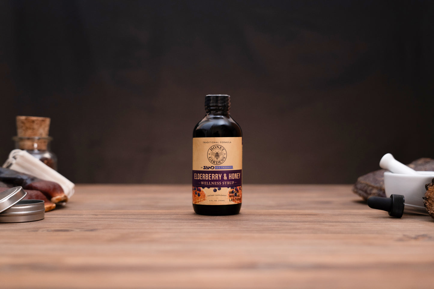 A small amber glass bottle of Zero Waste Labs Blueberry & Honey Infused Oil stands centered on a wooden tabletop against a dark, moody background. The bottle features a tan and purple label with a black cap, flanked by blurred rustic ingredients and containers on either side.