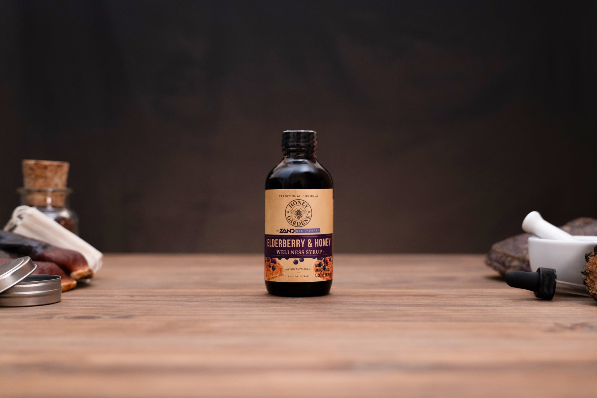 A small amber glass bottle of Zero Waste Labs Blueberry & Honey Infused Oil stands centered on a wooden tabletop against a dark, moody background. The bottle features a tan and purple label with a black cap, flanked by blurred rustic ingredients and containers on either side.