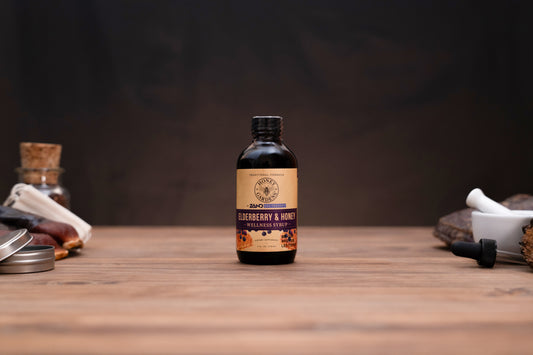 A small amber glass bottle of Zero Waste Labs Blueberry & Honey Infused Oil stands centered on a wooden tabletop against a dark, moody background. The bottle features a tan and purple label with a black cap, flanked by blurred rustic ingredients and containers on either side.