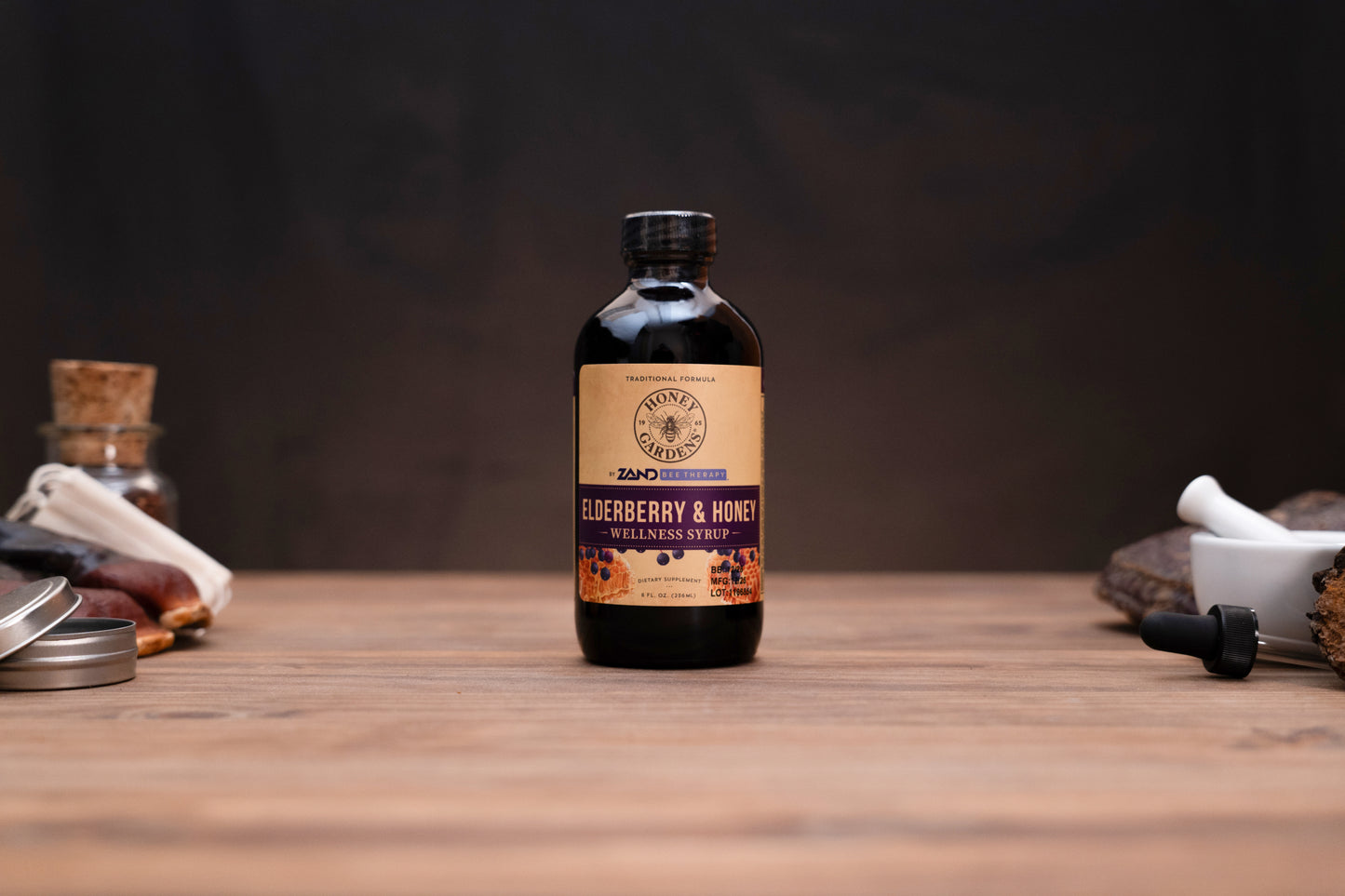 A bottle of Zero Waste Apothecary Elderberry & Honey Organic Syrup stands on a wooden tabletop against a dark background, flanked by rustic props including a small glass jar with a cork stopper, a white mortar and pestle, and a metal dropper.
