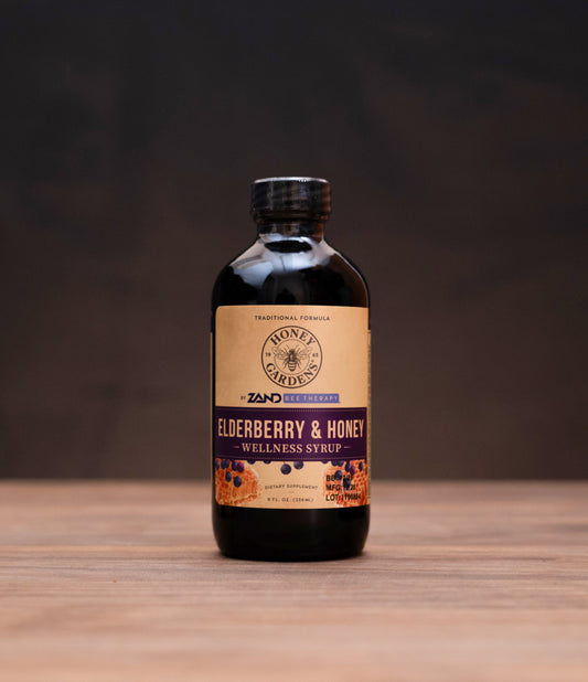 A bottle of Zero Waste Apothecary Elderberry & Honey Organic Syrup stands on a wooden tabletop against a dark background, flanked by rustic props including a small glass jar with a cork stopper, a white mortar and pestle, and a metal dropper.