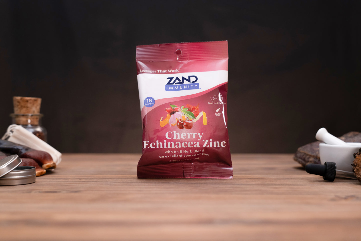 A bag of Zand Immunity Cherry Echinacea Zinc herbal supplement standing on a wooden surface against a dark background, with small blurred props on both sides.