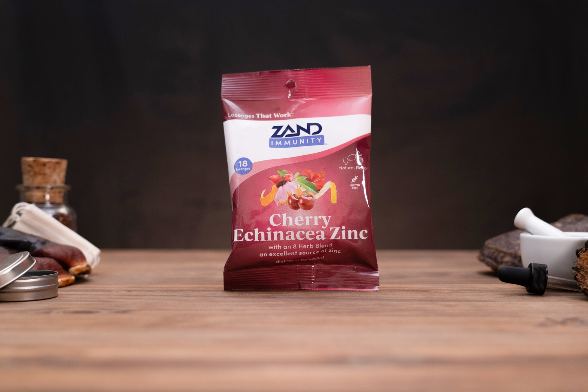 A bag of Zand Immunity Cherry Echinacea Zinc herbal supplement standing on a wooden surface against a dark background, with small blurred props on both sides.