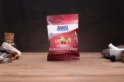 A bag of Zand Immunity Cherry Echinacea Zinc herbal supplement standing on a wooden surface against a dark background, with small blurred props on both sides.