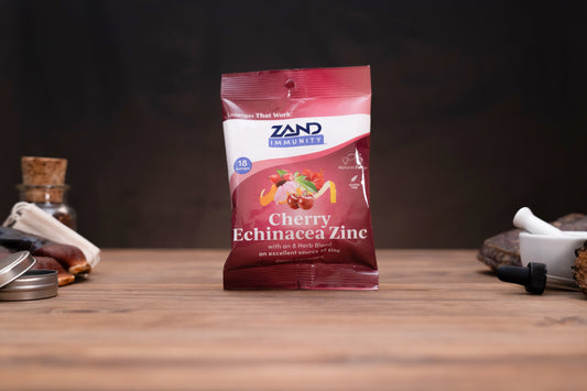 A bag of Zand Immunity Cherry Echinacea Zinc herbal supplement standing on a wooden surface against a dark background, with small blurred props on both sides.