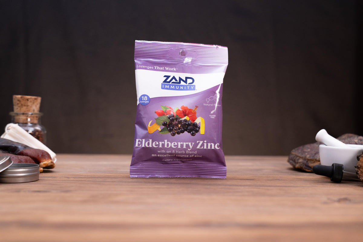 A purple and white bag of Zand Immunity Elderberry Zinc supplement stands on a wooden surface against a dark background, with small blurred herbal and laboratory props on either side.