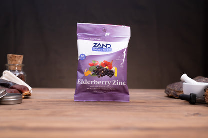 A purple and white bag of Zand Immunity Elderberry Zinc supplement stands on a wooden surface against a dark background, with small blurred herbal and laboratory props on either side.