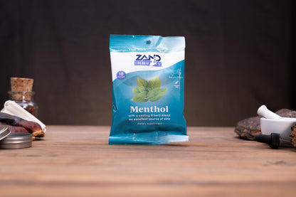 A blue and white packet of Zand The Market Menthol tincture standing on a wooden surface against a dark background, with small rustic props visible on both sides.