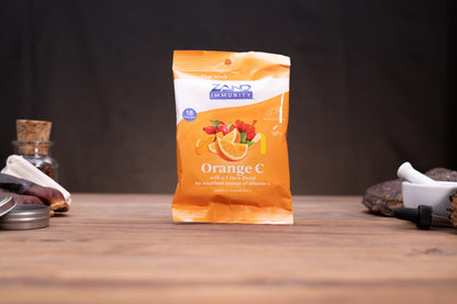 A stand-up pouch of Zand Immunity Orange C supplement standing on a wooden surface against a dark background, with small blurred props on both sides.